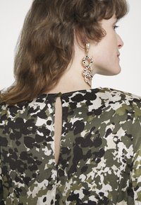 Tecido estampado floral em verde, preto e branco; apresenta um detalhe nas costas em forma de chave e um grande brinco em forma de serpente com acentos dourados.