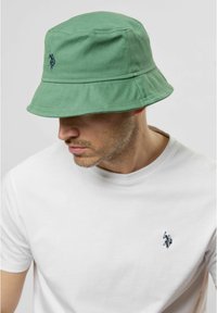 Grön bucket hat i bomull, med en bred brat och en blå broderad logotyp. Bärs med en enkel vit t-shirt.