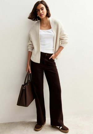 Femme portant un cardigan en tricot crème, un débardeur blanc, un pantalon large marron foncé, des chaussures marron à enfiler, tenant un grand sac à main marron foncé.