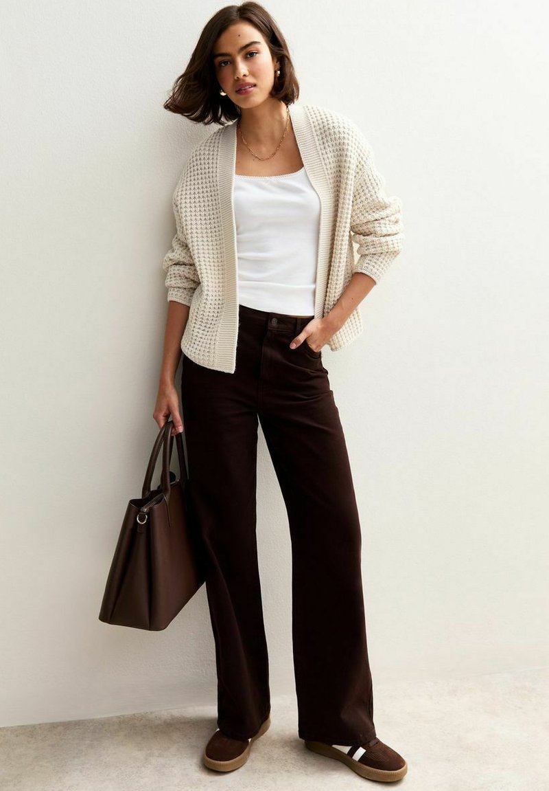 Femme portant un cardigan en tricot crème, un débardeur blanc, un pantalon large marron foncé, des chaussures marron à enfiler, tenant un grand sac à main marron foncé.