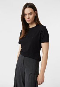 Kobieta z długimi brązowymi włosami, ubrana w czarną bluzkę z krótkim rękawem z napisem "NYC" oraz szare szerokie spodnie z wysokim stanem, stojąca na białym tle.