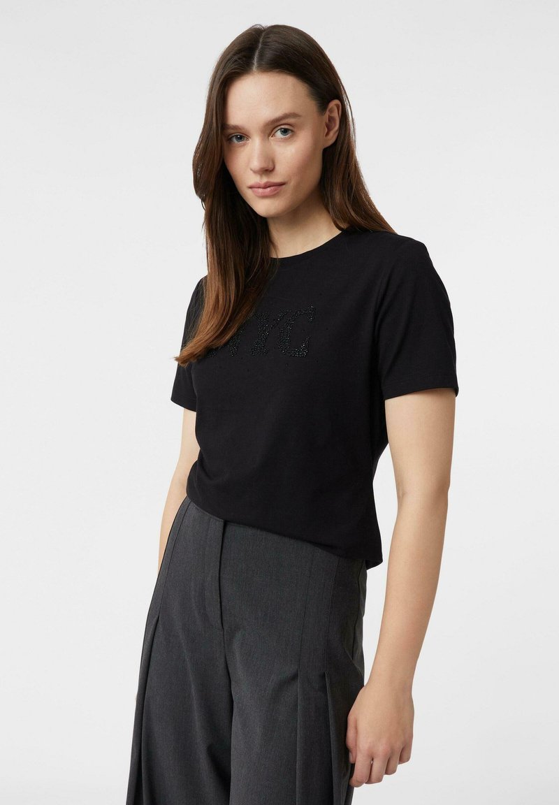 Kobieta z długimi brązowymi włosami, ubrana w czarną bluzkę z krótkim rękawem z napisem "NYC" oraz szare szerokie spodnie z wysokim stanem, stojąca na białym tle.