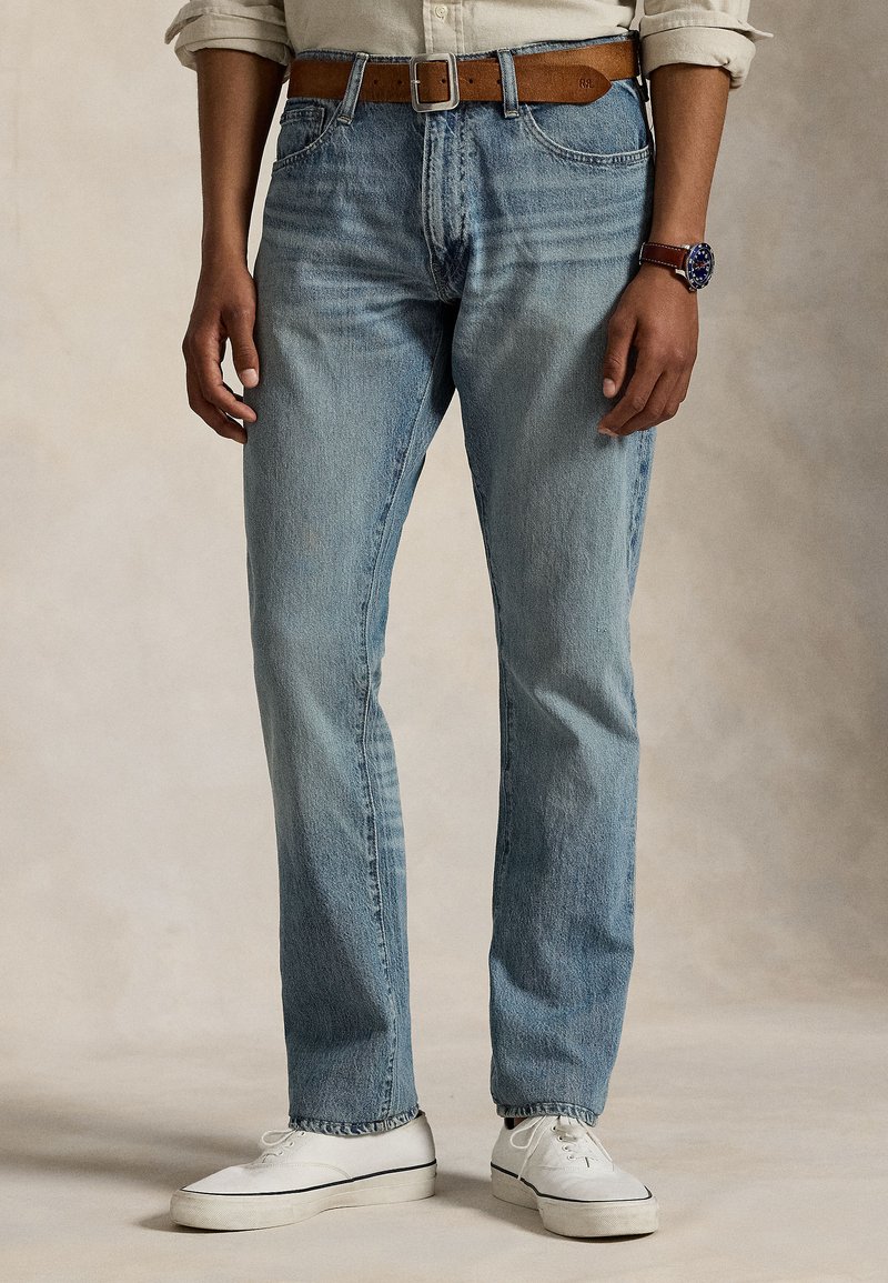 Calças de ganga azul claro com corte de perna direita, apresentando passadeiras para cinto e desbotamento subtil, combinadas com sapatilhas brancas e um cinto de couro castanho.