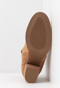 Tan perforované boty s hnědou gumovou podrážkou, blokovým podpatkem a bočním zipem. Design obsahuje texturovaný povrch a kruhové vzory.