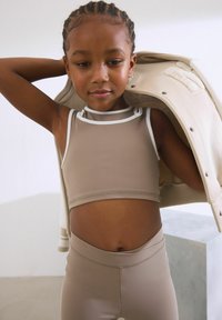 Brun sportcrop-top med vit kant och matchande högmidjade leggings. En beige jacka hänger över ena axeln och visar ett slätt tyg.
