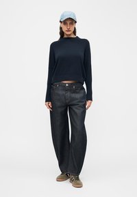 Femme portant une casquette bleu clair, un pull bleu marine foncé, un jean noir à jambes larges et des baskets vert olive, debout devant un fond blanc uni.