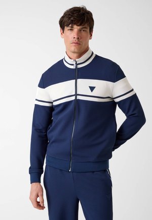 Mann steht mit der Hand hinter dem Rücken, trägt eine marineblaue und weiße Trainingsjacke mit Reißverschluss und passende Hose vor einfachem weißem Hintergrund.