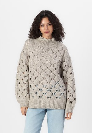 Beige strikkegenser med høy krage, åpen diamantmønster og ribbestrikkede mansjetter. Kombinert med lyseblå jeans.
