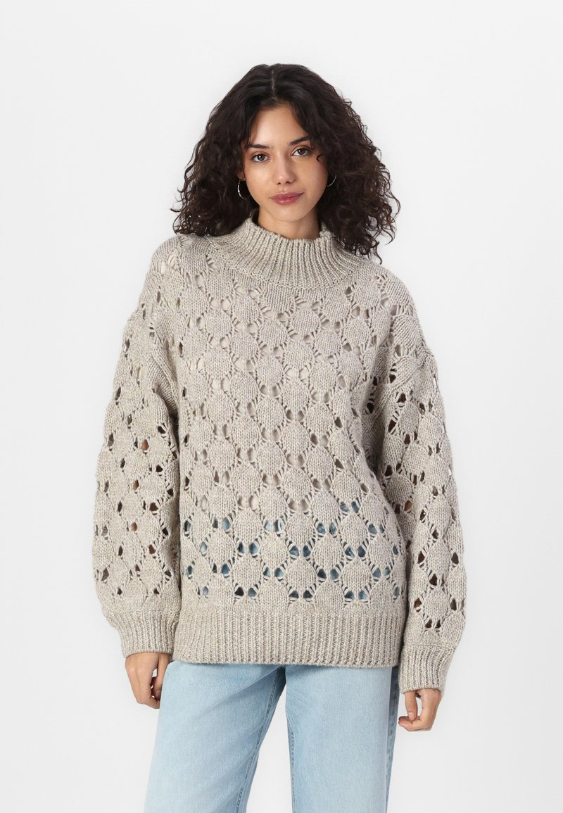Beige strikkegenser med høy krage, åpen diamantmønster og ribbestrikkede mansjetter. Kombinert med lyseblå jeans.