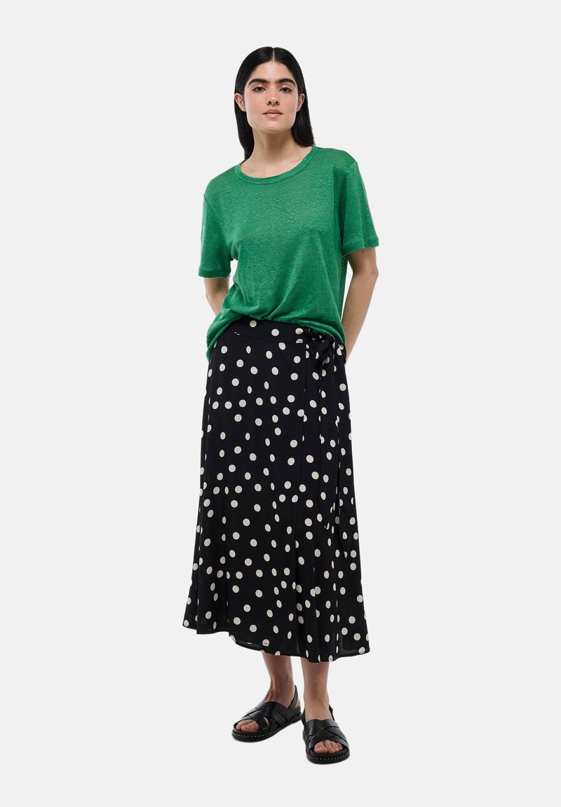 Haut vert à manches courtes associé à une jupe portefeuille noire à pois. Matériaux texturés, coupe décontractée et sandales plates noires complètent le look.