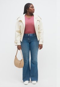 Γυναίκα με κοτσιδωτά μαλλιά, φοράει εκρού μπουφάν, βιολετί μπλούζα, μπλε τζιν, λευκά αθλητικά παπούτσια, κρατάει μπεζ τσάντα ώμου, κοιτώντας προς τα πλάγια.