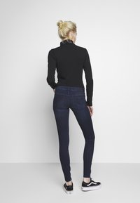Top negro de manga larga combinado con jeans skinny azul oscuro, que presentan una textura suave, una cintura de altura media y un diseño clásico de cinco bolsillos. Zapatillas negras completan el look.