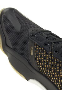 Zapatilla deportiva negra con parte superior de malla, panel lateral texturizado y detalle dorado "STABIL". Suela de goma con una entresuela clara en contraste.