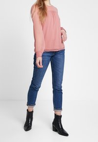Blusa trasparente rosa chiaro con maniche lunghe e polsini arricciati, abbinata a jeans di denim scuri e stivaletti neri con tacco a blocco.
