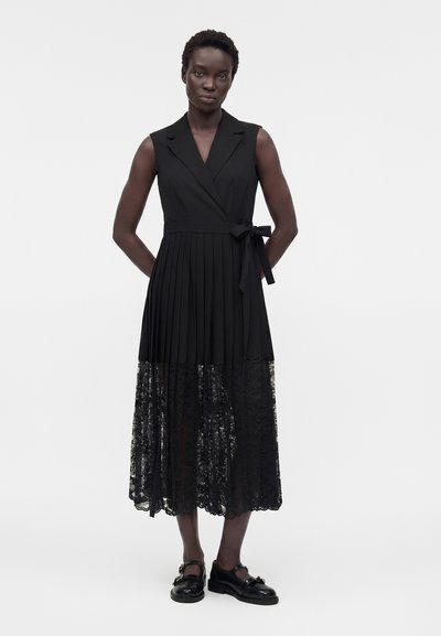 Γυναίκα που στέκεται με τα χέρια πίσω από την πλάτη φορά αμάνικο μαύρο φόρεμα με πλισέ φούστα και δαντελένιο τελείωμα, συνδυασμένο με μαύρα παπούτσια.