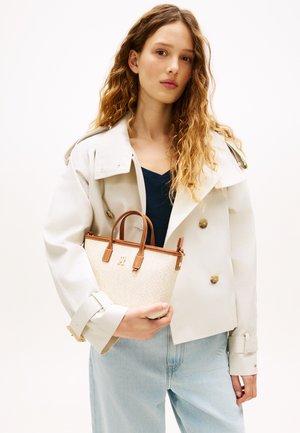 Νεαρή γυναίκα με μακριά σγουρά μαλλιά φοράει μπεζ καμπαρντίνα και μπλε τζιν, κρατώντας μικρή μπεζ τσάντα με καφέ λαβές στο ύψος της μέσης.