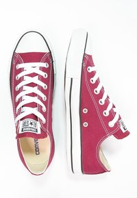 Tmavě červené plátěné tenisky s bílými tkaničkami a gumovou podrážkou. Mají černé pruhy, kovové dírky a klasický nízký střih.