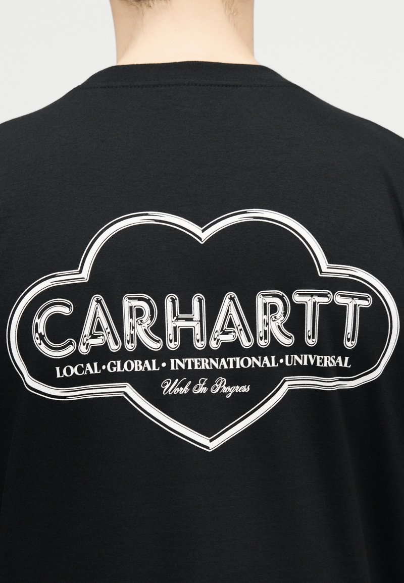 Persona che indossa una maglietta nera con un logo a forma di cuore contornato di bianco con la scritta "Carhartt Local Global International Universal Work In Progress" sulla schiena.