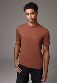 Tricou scurt cu mânecă închisă, de culoare maro, cu un design ajustat și decolteu rotund. Textură netedă, stil minimalist, asortat cu blugi negri.