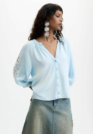 Vrouw met lang donker haar met grote witte bloemvormige oorbellen, een lichtblauw gebuttond bloesje met geborduurde mouwen en een spijkerrok.
