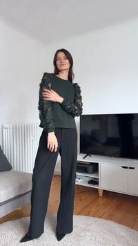 Temno zelena majica z teksturiranimi prosojnimi cvetličnimi rokavi, kombinirana s črnimi širokimi hlačami in črnimi koničastimi petami. Vidni preproga in pohištvo.