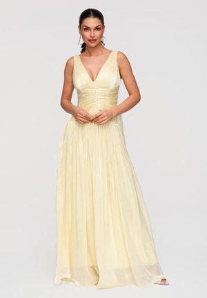 Mujer con un vestido de noche sin mangas, amarillo pálido y plisado, con un escote en V profundo, de pie con las manos suavemente entrelazadas.