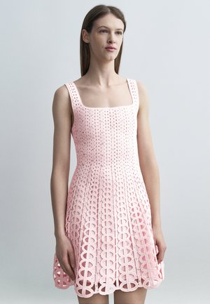Žena má na sebe ružové háčkované šaty bez rukávov so štvorcovým výstrihom a kruhovými otvorenými vzormi na sukni.