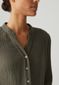 Olive green, teksturowana, koszula zapinana na guziki z kołnierzykiem mandaryńskim, z subtelnymi pionowymi liniami i guzikami przypominającymi perły.