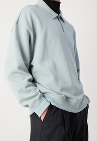 Ljusblå tröja med zip-krage, ribbade ärmslut och nederkant. Slätt material, avslappnad passform och diskreta ränder på ärmsluten.