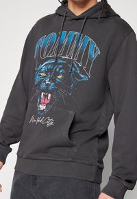 Mustat huppari, jossa on graafinen kuva murisevasta mustasta pantterista ja "TOMMY" sinisellä tekstillä. Varustettu kenguru-taskulla ja ribbaatuilla hihoilla.