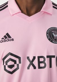 Vaaleanpunainen jalkapallopaita, joka on valmistettu sileästä kankaasta, mustilla yksityiskohdilla, kauluksella sekä Adidas- ja Club Internacional de Miami -logolla.