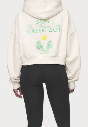 Person trägt einen kurzen cremefarbenen Hoodie mit grünem Text "SUN The Wholesome Life CAME OUT" und einem Sonnen- sowie Palmblätter-Design auf der Rückseite.