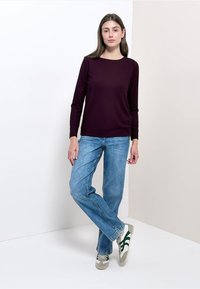 Maglietta a maniche lunghe color amaranto con collo rotondo, abbinata a jeans a gamba dritta azzurri chiari e sneakers con dettagli verdi su sfondo neutro.