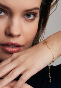 Złoty bransoletka łańcuszkowa z zawieszką w kształcie kwiatu i zielonym koralikiem akcentującym. Łańcuszek jest delikatny, z regulowanym zapięciem i subtelną fakturą.