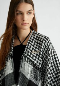 Sciarpa houndstooth bianca e nera con bordi frangiati, decorata con un embellishment metallico, indossata sopra un top nero, creando un aspetto testurizzato.
