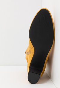 Botte en daim jaune positionnée semelle vers le haut contre un mur d'angle blanc, montrant une semelle noire texturée et un détail de fermeture éclair latérale.