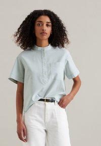 Camicetta azzurra a righe con maniche corte, colletto alla cinese, pattina dei bottoni e taschino frontale, abbinata a pantaloni bianchi e una cintura nera.