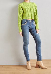 Suéter corto de punto trenzado verde brillante con mangas largas, combinado con jeans ajustados azules y botas tobilleras de imitación de cocodrilo beige.