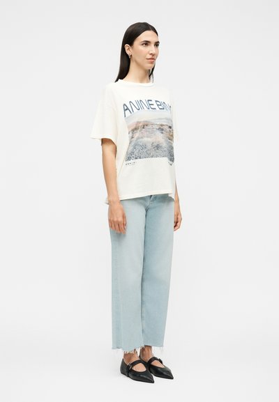 Γυναίκα στέκεται φορώντας λευκό oversized μπλουζάκι με γραφικό, ανοιχτό μπλε τζιν με ίσια γραμμή και ξεφτισμένα τελειώματα, και μαύρες μυτερές μπαλαρίνες με λουράκια.