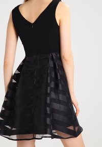 Svart klänning med en figurnära överdel och V-bak, lager med en genomskinlig kjol som har horisontella satinstreck och en dold dragkedja.