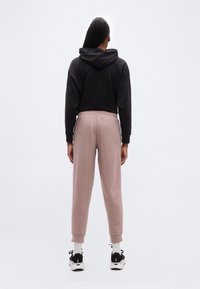 Άτομο που φοράει μαύρο φούτερ με κουκούλα, ροζ-σκουρόχρωμο παντελόνι φόρμας, λευκές κάλτσες και μαύρα αθλητικά παπούτσια, στέκεται με την πλάτη προς το μέρος της κάμερας μπροστά σε ένα απλό φωτεινό φόντο.