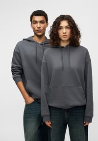 Σκούρες γκρι φούτερ με κουκούλα από μαλακό ύφασμα, με μπροστινές τσέπες και κορδόνια, φορεμένα με φαρδιές τζιν παντελόνες.