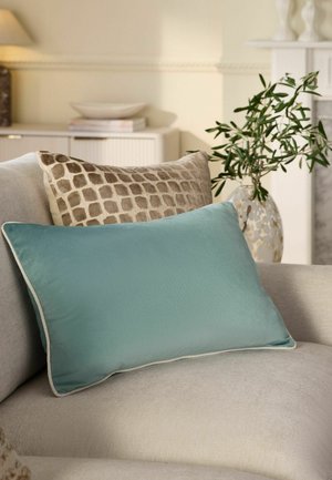 Beiges Sofa mit einem blauen rechteckigen Kissen vorne und einem gemusterten quadratischen Kissen dahinter, neben einer Vase mit grünen blättrigen Zweigen.