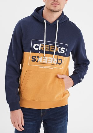 Hoodie met een marineblauwe bovenkant en mosterdgele onderkant, voorzien van een kangoeroezak, witte trekkoorden en een grafische logo op de voorkant.