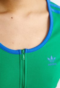 Zelený vroubkovaný crop top s modrým límečkem, který má přední zip se zeleným pullerem a malým modrým logem Adidas na prsou.