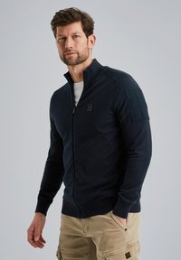 Navy zip-up sweater with a high collar, textured knit, and patch pocket on the left sleeve, paired with light khaki cargo pants.