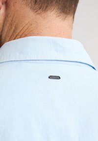 Cămașă de un albastru deschis, dintr-un material texturat, cu guler. O etichetă metallică mică cu "Paredes" este atașată pe spate, aproape de gât.