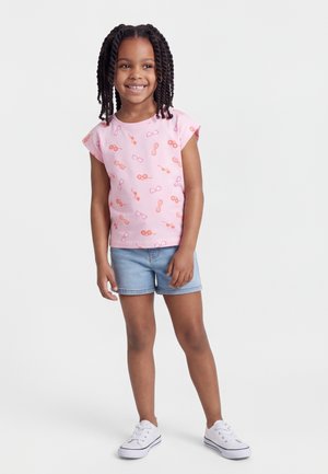 Χαμογελαστή κοπέλα με πλεγμένα μαλλιά που φοράει ροζ μπλουζάκι με στάμπα γυαλιών ηλίου, ανοιχτόχρωμα τζιν σορτς και λευκά αθλητικά παπούτσια, στέκεται σε λευκό φόντο.