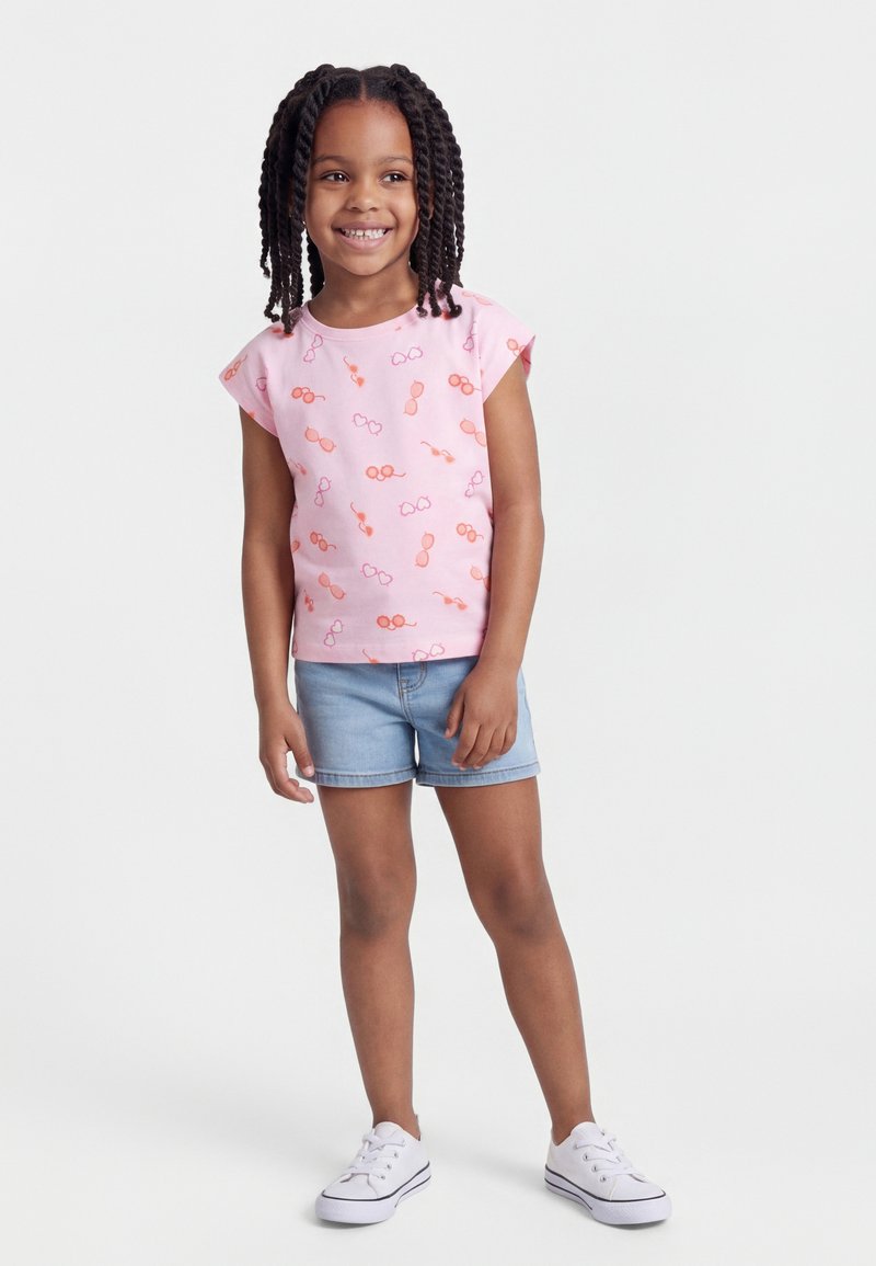 Lächendes Mädchen mit geflochtenem Haar, das ein pinkfarbenes Oberteil mit Sonnenbrillenmuster, helle Jeansshorts und weiße Turnschuhe trägt und auf weißem Hintergrund steht.