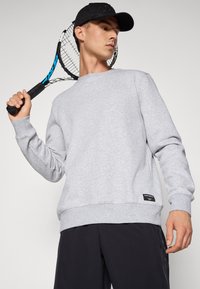 Felpa grigia con maniche lunghe, polsini e orlo a coste, tessuto testurizzato, indossata con un cappellino nero, mentre tiene una racchetta da tennis in una posa dinamica.